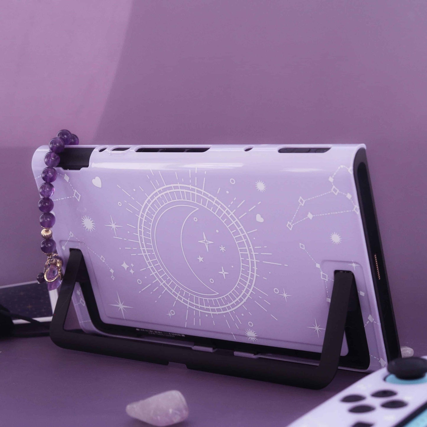 Cosmic Moon Nintendo Switch 2 case shown from the back with kickstand propped up, moon and constellation print on lilac, with an amethyst bracelet and crystal nearby on a purple set.