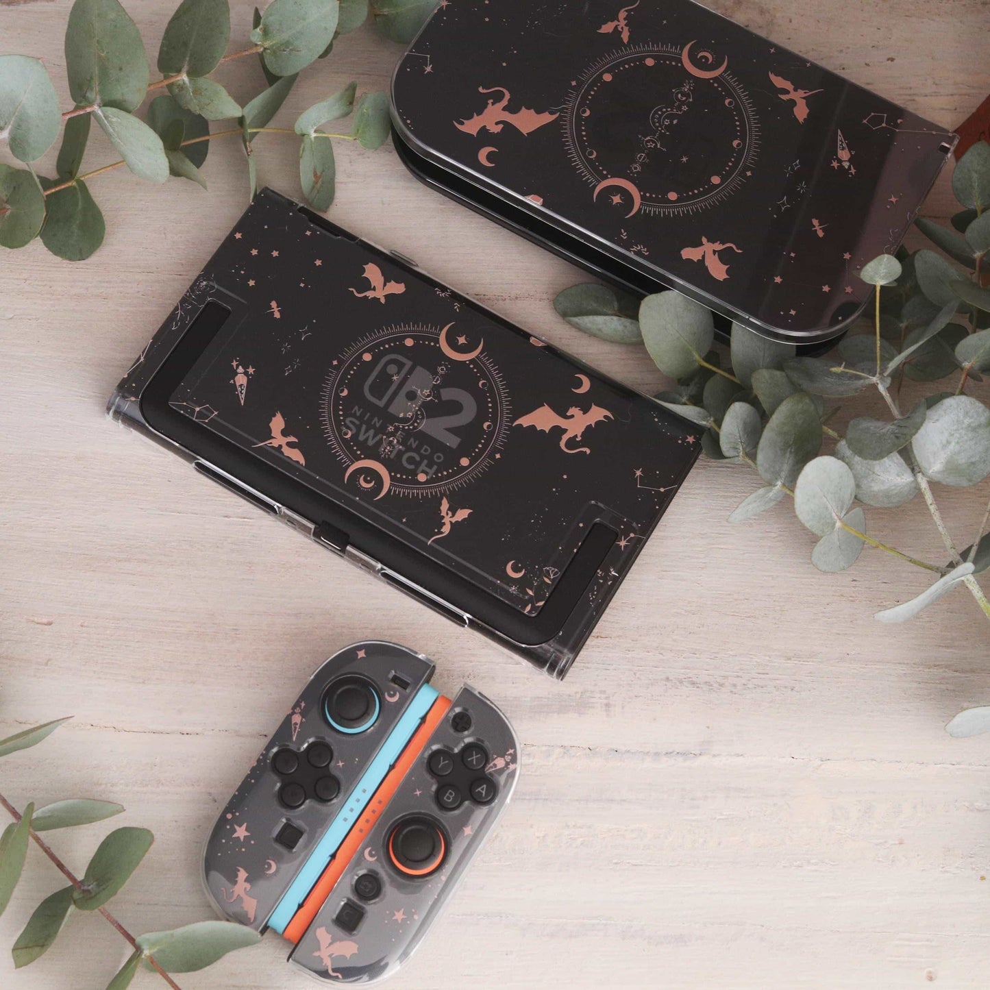 Emberwing Nintendo Switch 2 case, dock plate, and Joy-Cons laid out together showing the matching dragon, moon, and star design, on a wooden surface with eucalyptus leaves.