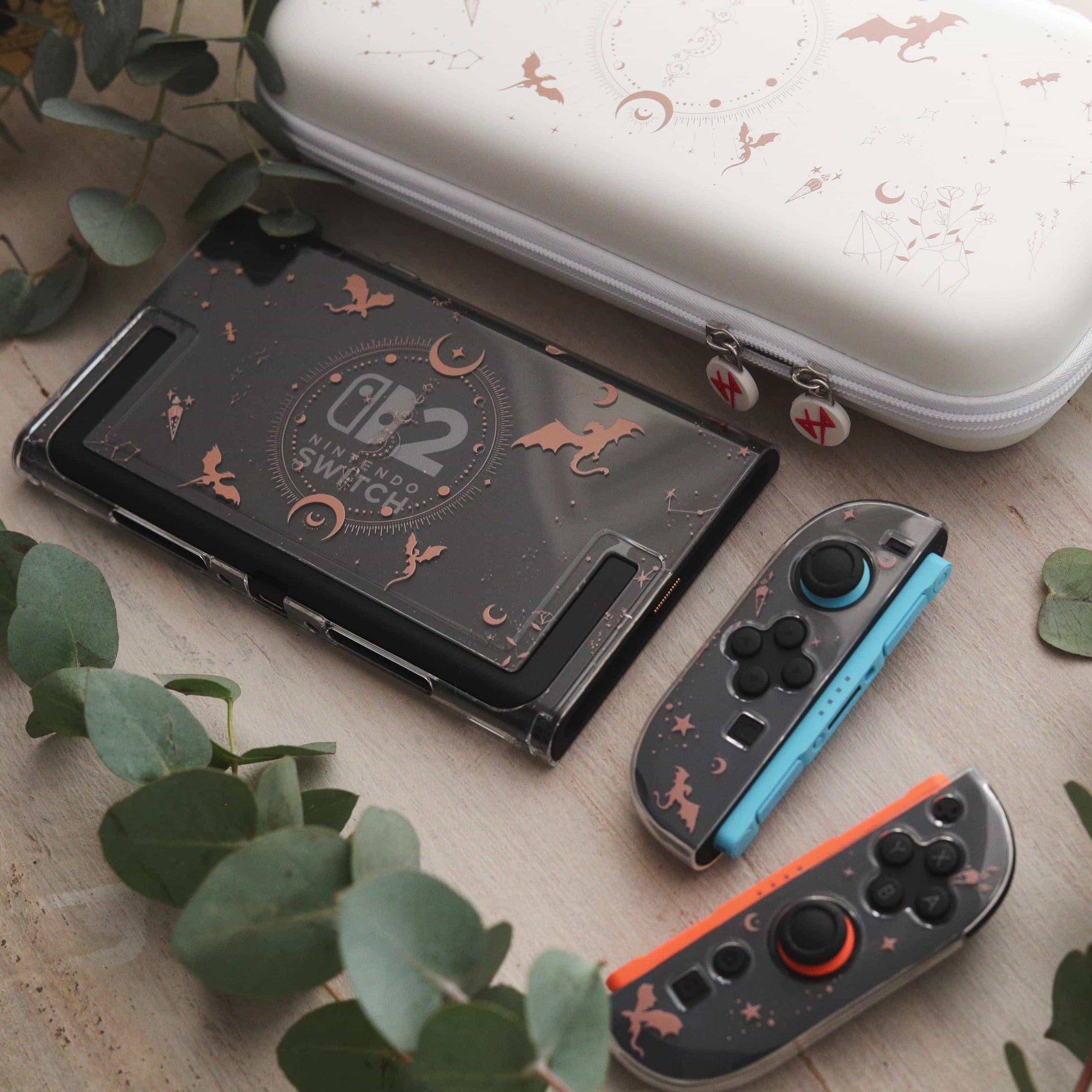 Emberwing Nintendo Switch 2 console with detached blue and orange Joy-Cons, dragon and moon designs visible, alongside the matching carry case and eucalyptus leaves.