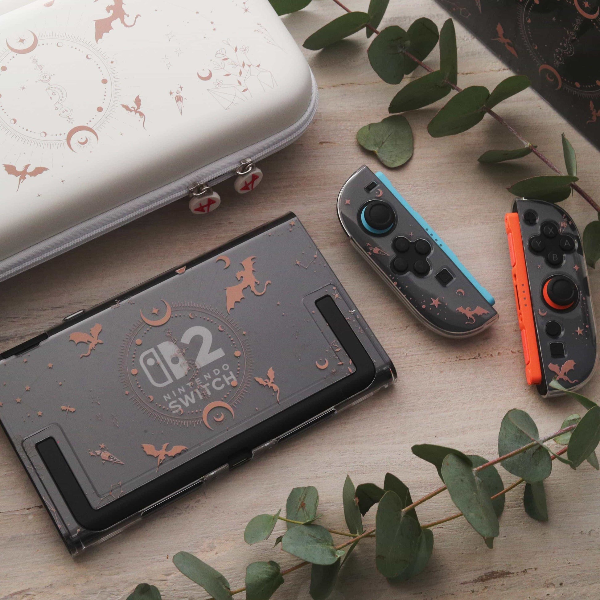 Emberwing Nintendo Switch 2 case with detached Joy-Cons in blue and orange, dragon and moon print visible, set beside the cream carry case on a eucalyptus-covered surface.