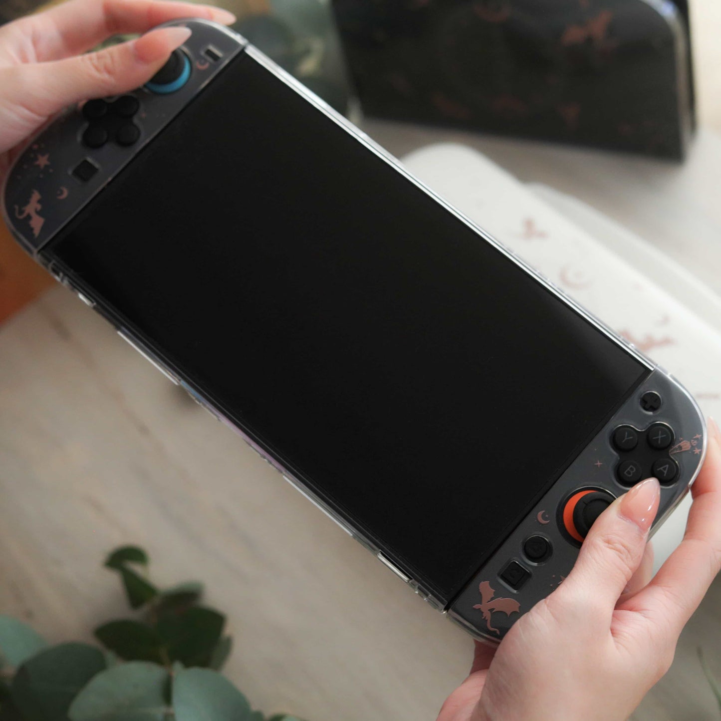 Hands holding a Nintendo Switch 2 fitted in the Emberwing case, dragon and moon details visible on the Joy-Cons, screen off, with the carry case and eucalyptus in the background.