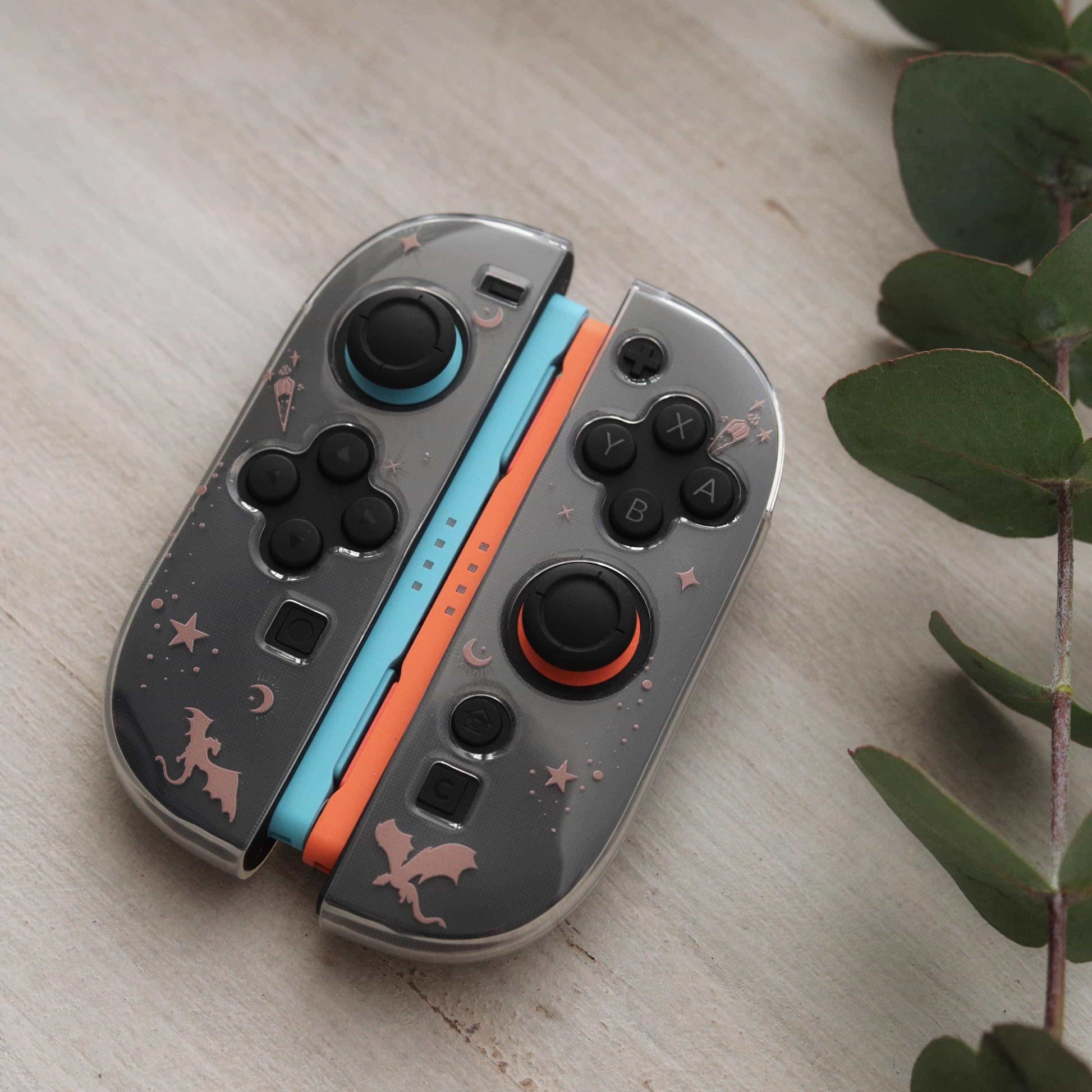 Emberwing Joy-Con controllers attached together, showing dragon, moon, and star design on grey shells with blue and orange accents, eucalyptus branch beside them.