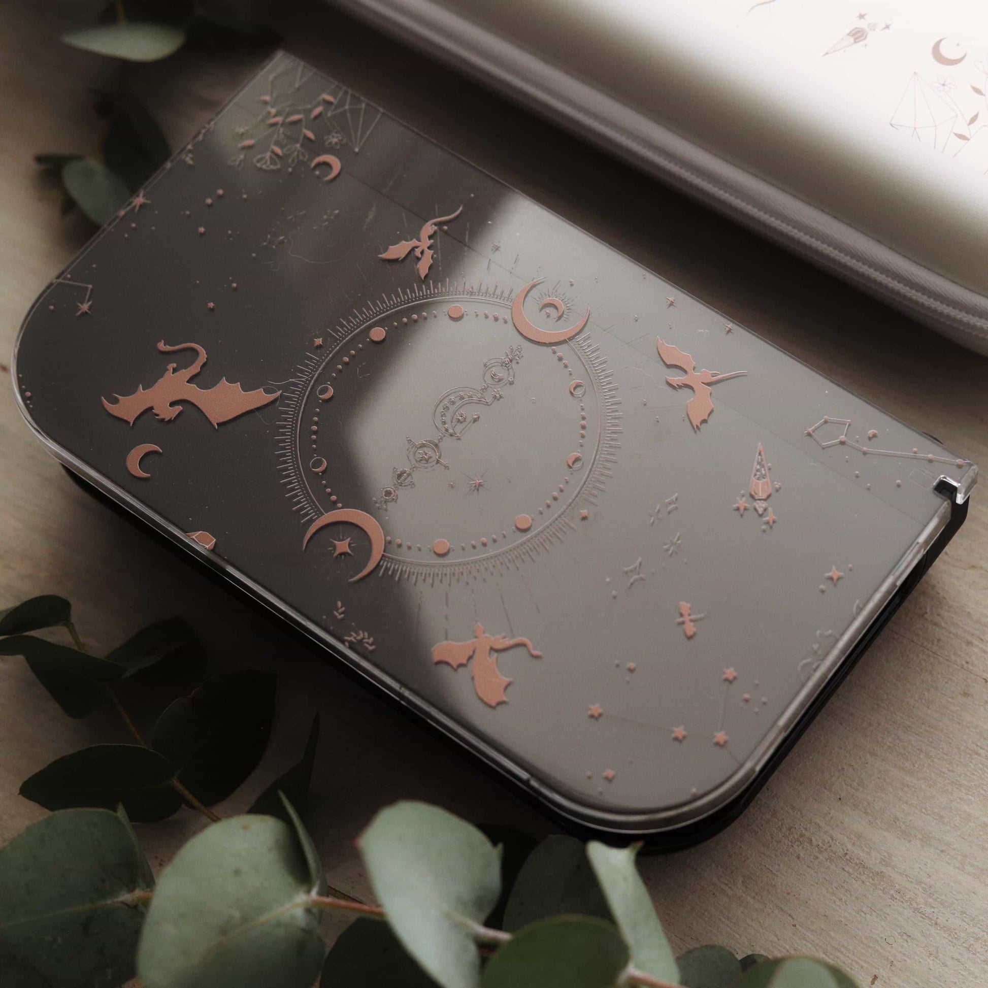 Emberwing Nintendo Switch 2 dock plate with engraved dragon, moon, and constellation design, resting on a light surface surrounded by eucalyptus leaves.