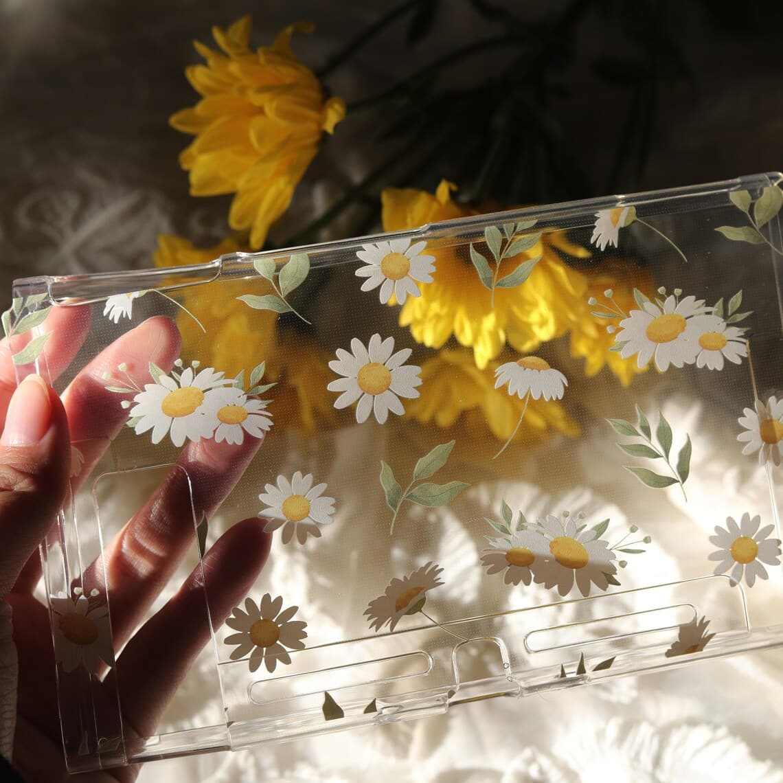 Hand holding clear Nintendo Switch Daisy Daydream console shell showing transparent daisy floral print with yellow flowers in background